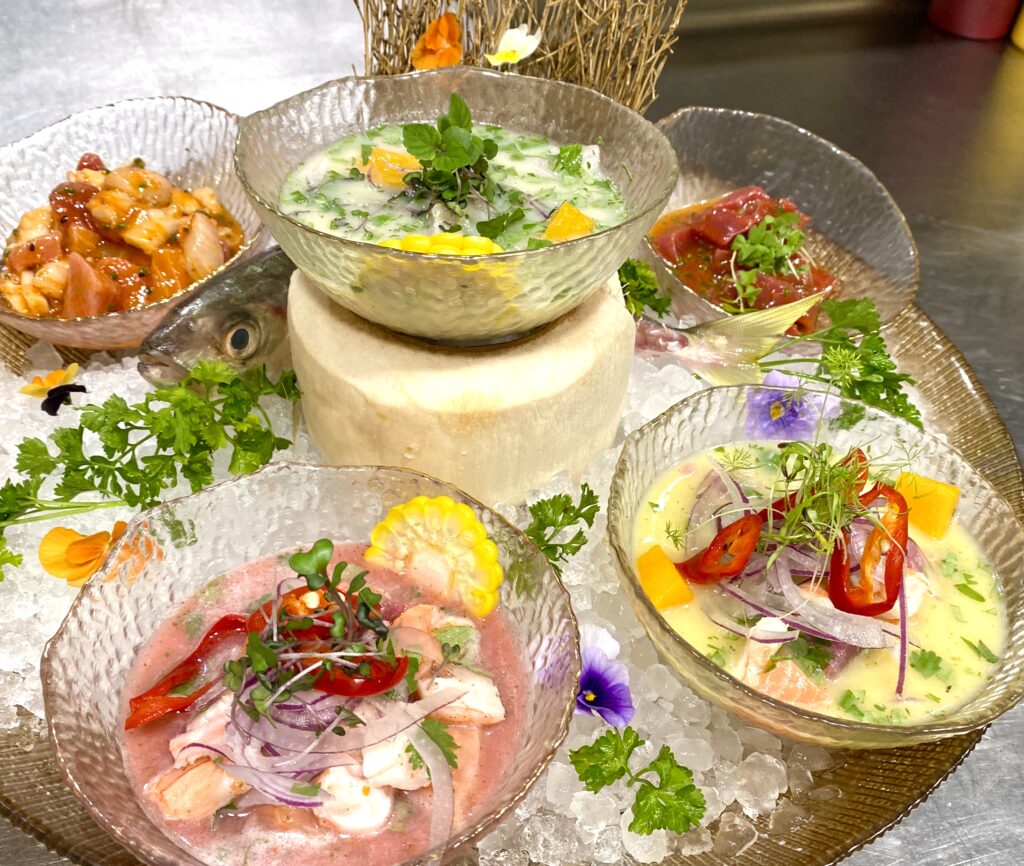 Varios platos de ceviche servidos en copas y cuencos, con cebolla morada, ají, maíz y flores comestibles